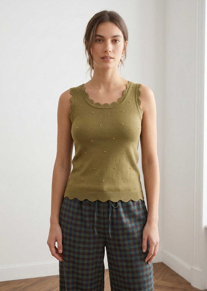 A woman modeling a sleeveless olive green knit top with scalloped edges and small bead details, paired with plaid pants, standing in a bright indoor room.
