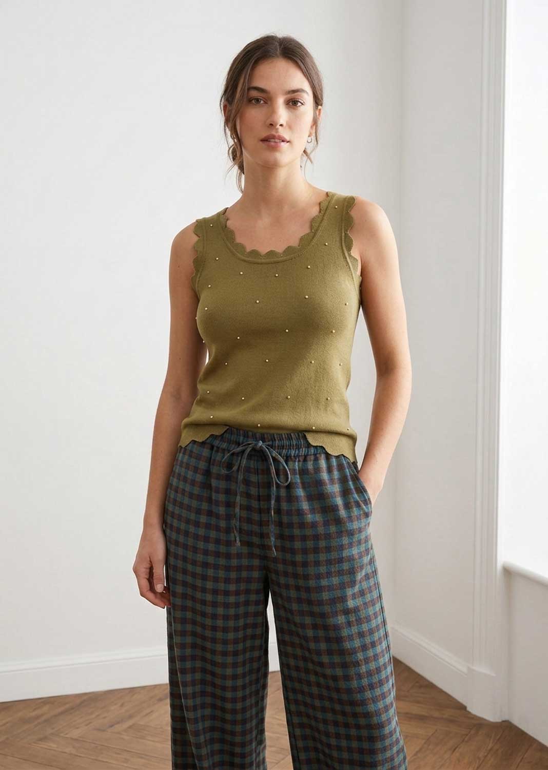 A woman modeling a sleeveless olive green knit top with scalloped edges and small bead details, paired with dark plaid drawstring pants, standing in a bright room with white walls and wood flooring.