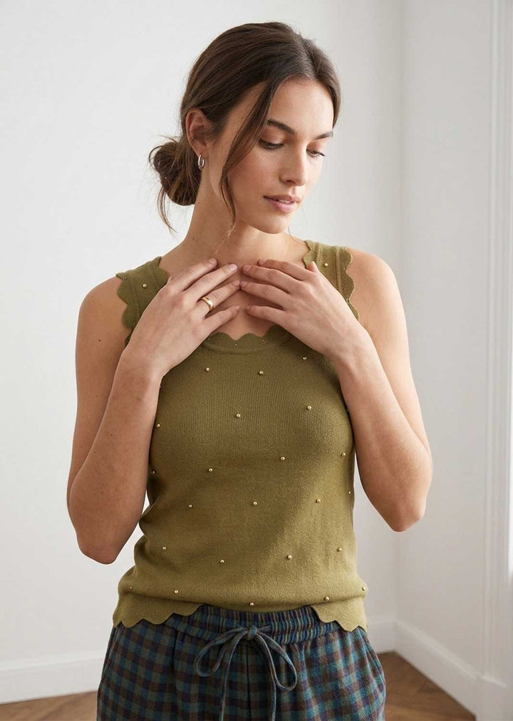 A woman wearing an olive green knit top with scalloped edges and small pearl-like embellishments, paired with plaid pants, standing in a bright indoor setting.