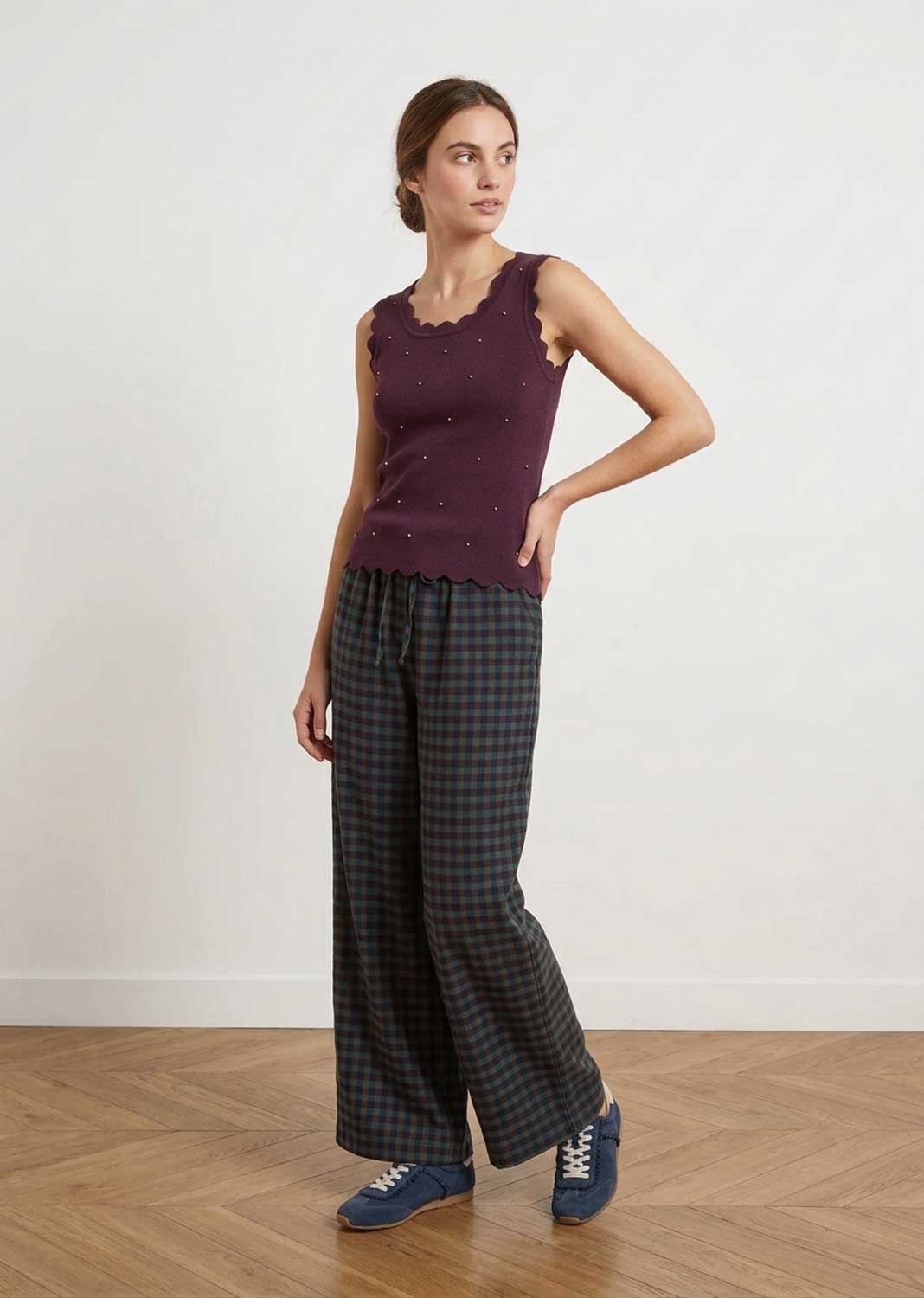 A woman modeling a sleeveless dark plum knit top with scalloped edges and small embroidered dots, paired with dark green and black plaid wide-leg trousers and blue sneakers, standing against a white wall on a wood floor.