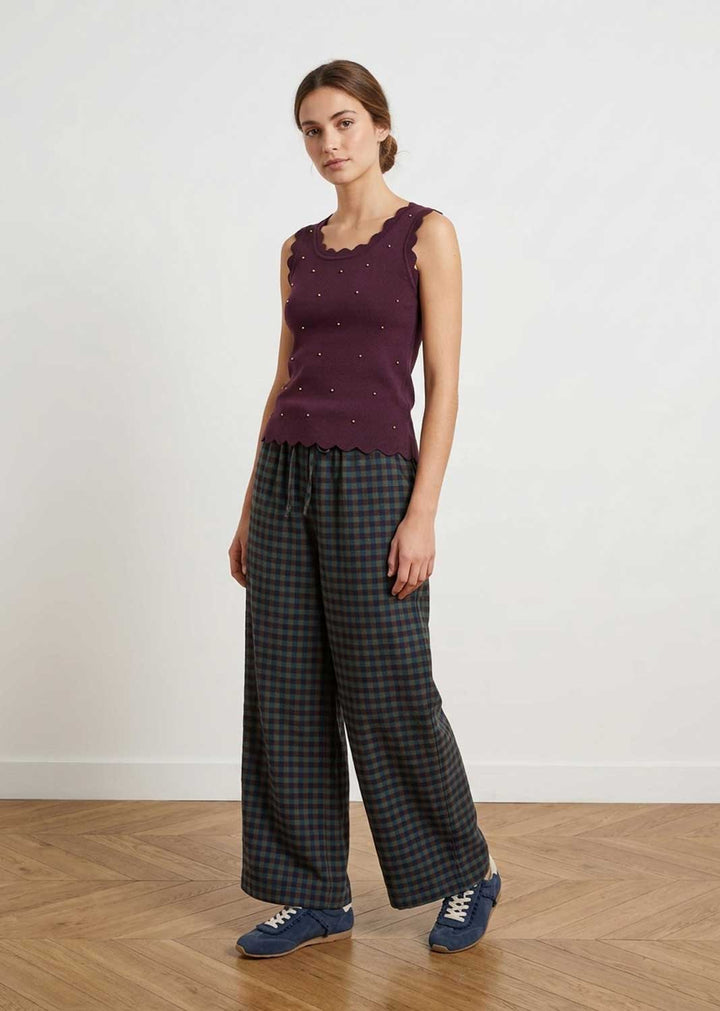 A woman stands in a studio wearing a sleeveless burgundy knit top with scalloped edges and small dot details, paired with dark green and black plaid wide-leg pants and blue sneakers.