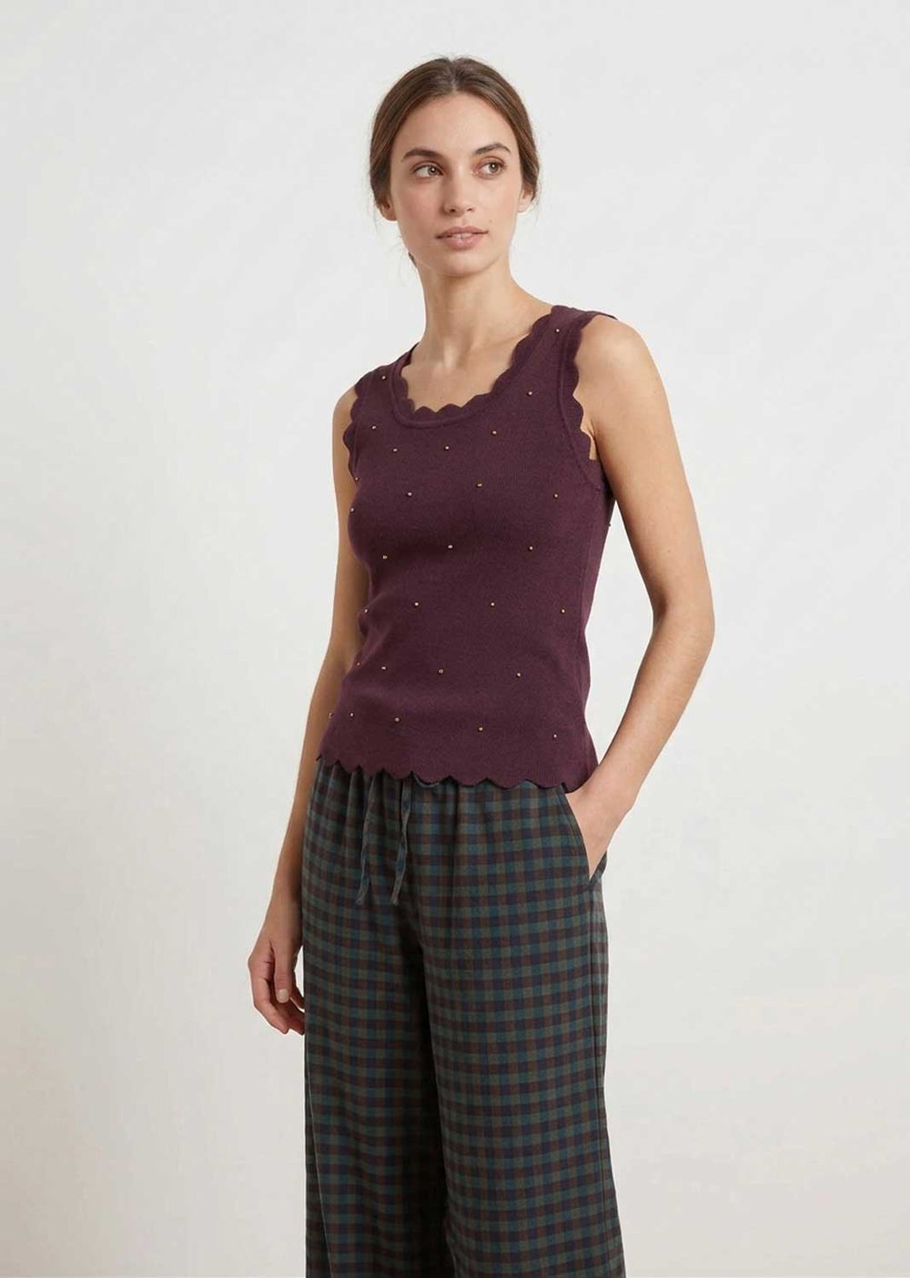 A woman modeling a sleeveless knit top in deep plum purple with scalloped edges and small embroidered details, paired with dark green and black plaid pants, against a plain light gray studio background.