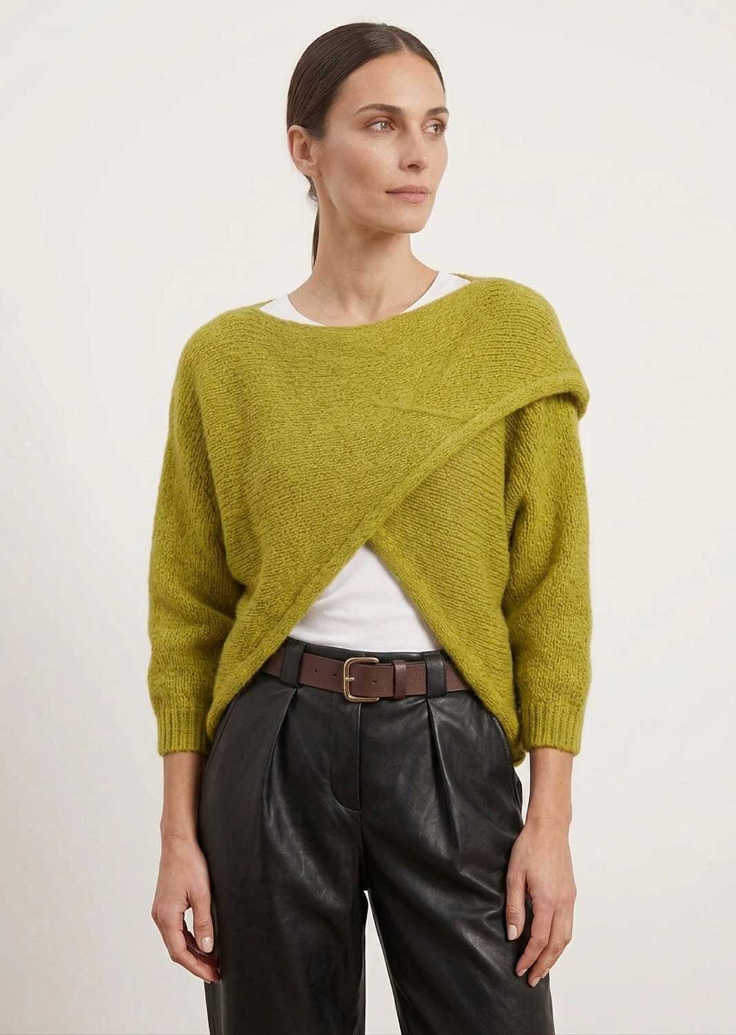 A woman modeling a textured olive green asymmetrical knit sweater over a white top and black leather pants, photographed against a plain light background.