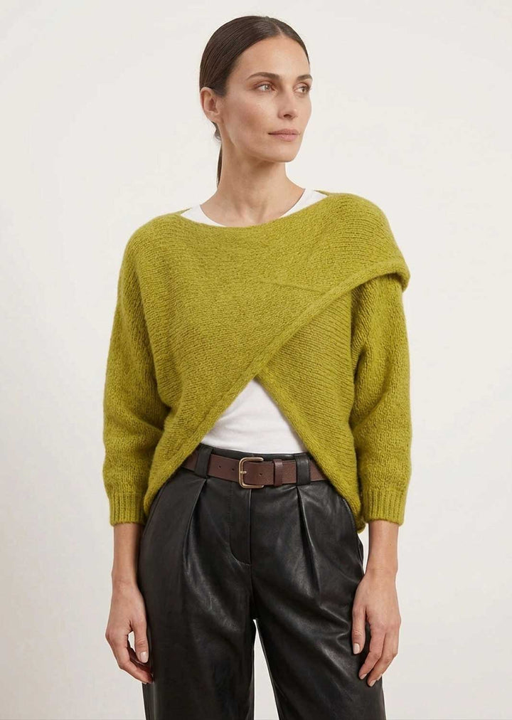 A woman modeling a textured olive green asymmetrical knit sweater over a white top and black leather pants, photographed against a plain light background.