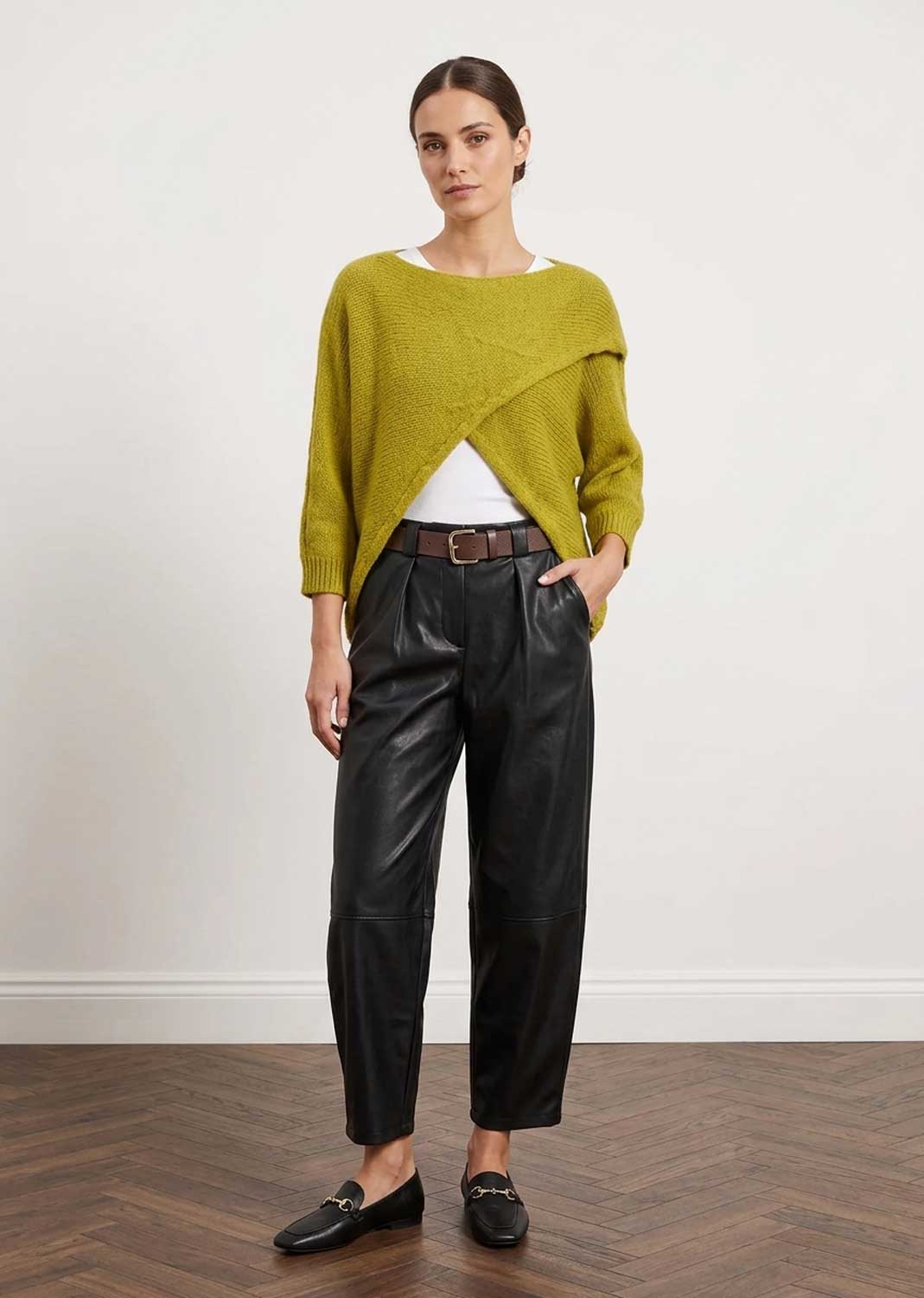 A woman stands in a studio wearing a textured olive green asymmetrical knit sweater, black leather trousers, a brown belt, and black loafers against a white wall and wood floor.