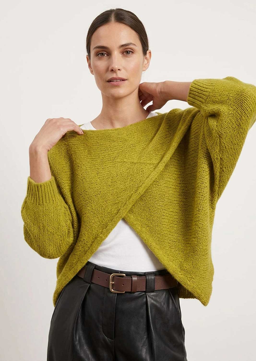 A woman modeling a textured olive green knit wrap-front sweater over a white top and black leather pants, photographed against a plain light background.