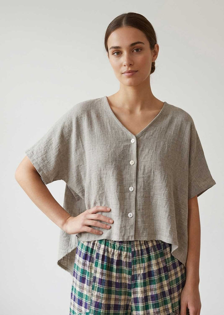 A woman modeling a light taupe linen short-sleeve button-up top with a V-neck, paired with green and blue plaid pants, against a plain light gray background.