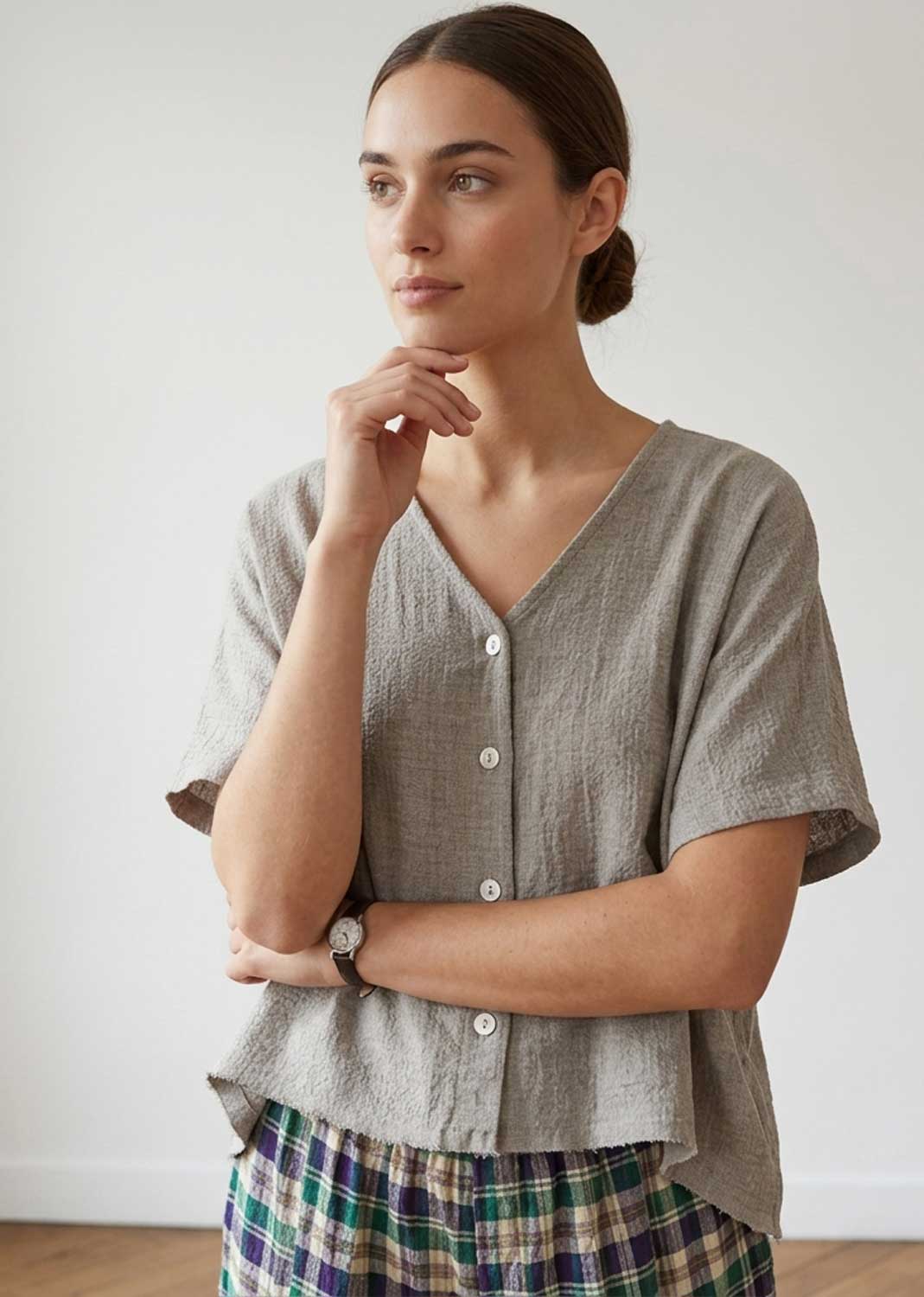 A woman wearing a light taupe linen short-sleeve button-up top and a green, blue, and white plaid skirt, standing against a plain white wall.