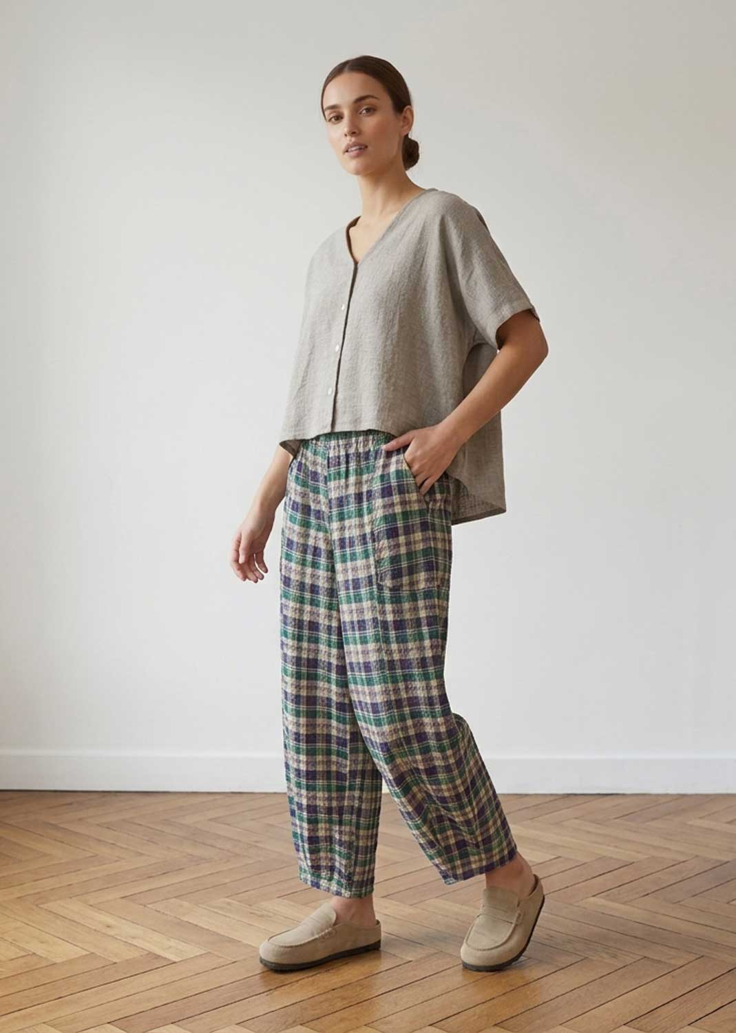 A woman modeling a light taupe short-sleeve cropped top and green, blue, and beige plaid trousers, standing on a wooden parquet floor against a white wall.