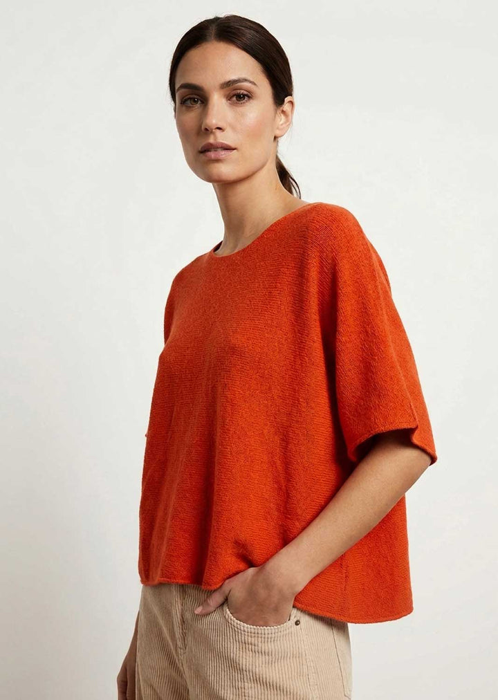 A woman wearing a bright orange short-sleeve knit top and beige corduroy trousers, photographed against a plain light gray studio background.