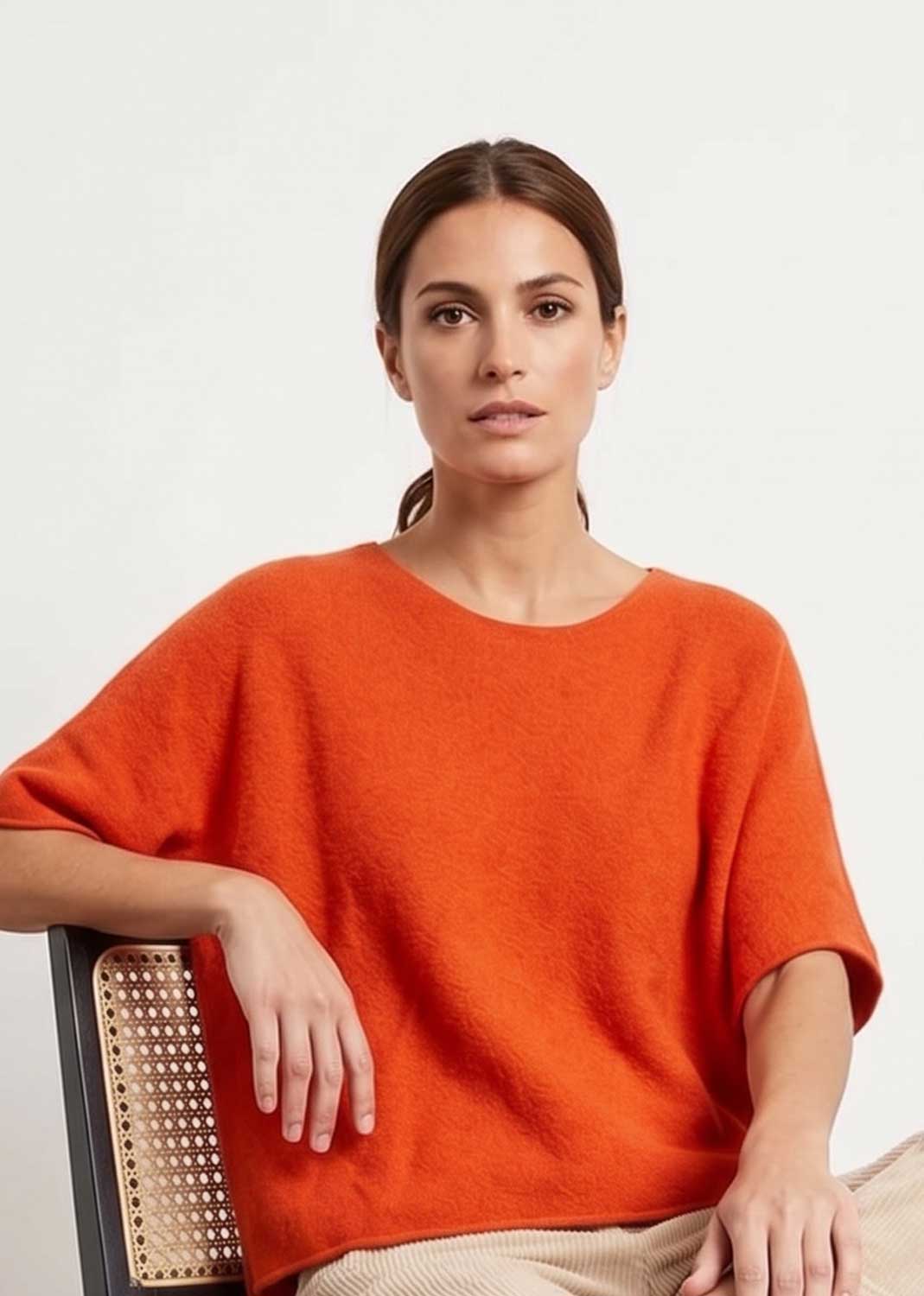 A woman wearing a bright orange short-sleeve knit top, seated on a chair with a woven back, against a plain light background.