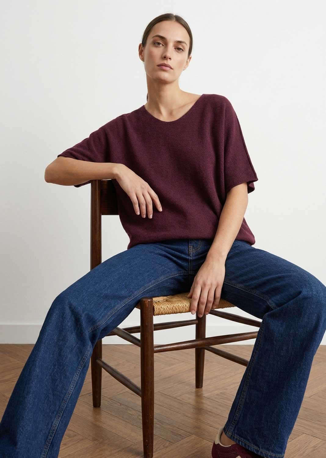A woman sits on a wooden chair wearing a loose, short-sleeve crew-neck top in deep burgundy and blue jeans, set against a minimalist white wall and light wood floor.