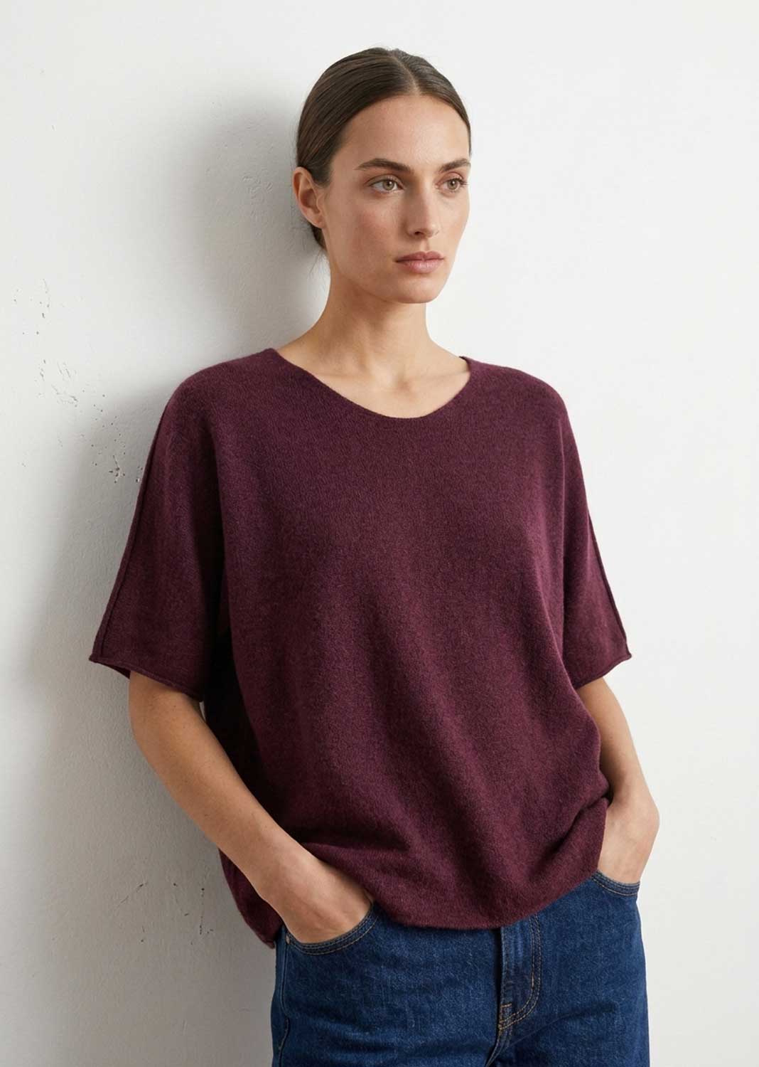 A woman wearing a short-sleeve crewneck knit top in heathered burgundy, paired with blue jeans, standing against a light off-white wall.