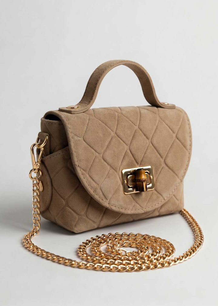 A small taupe croc suede handbag with a top handle, gold-tone clasp, and a long gold chain strap, photographed on a light gray background.