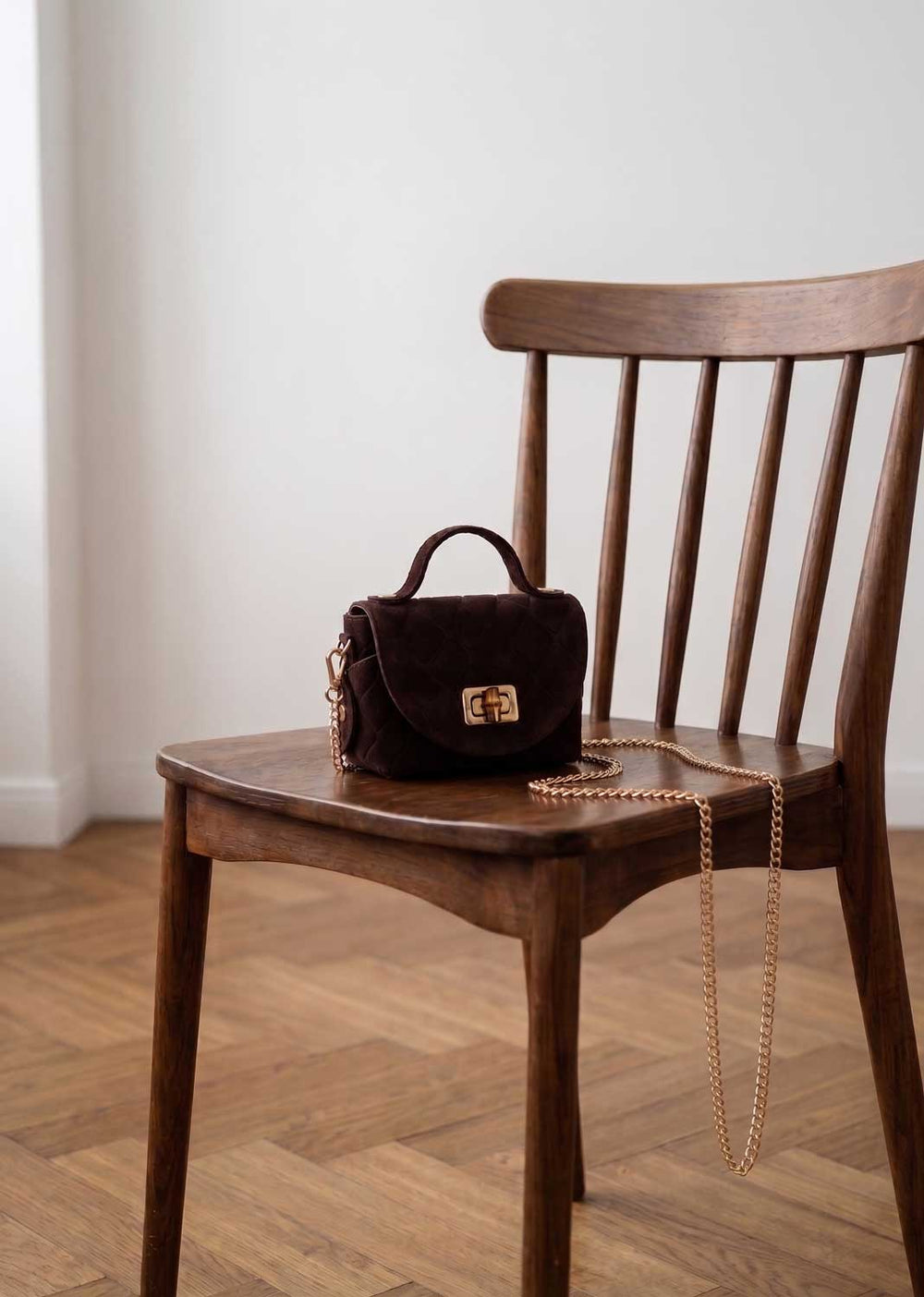 A dark brown croc suede handbag with a gold-tone turn-lock clasp and a gold chain strap, resting on a wooden spindle-back chair in a room with light-colored walls and a wood herringbone floor.