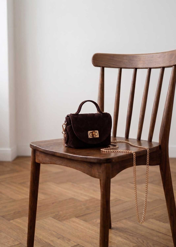 A dark brown croc suede handbag with a gold-tone turn-lock clasp and a gold chain strap, resting on a wooden spindle-back chair in a room with light-colored walls and a wood herringbone floor.