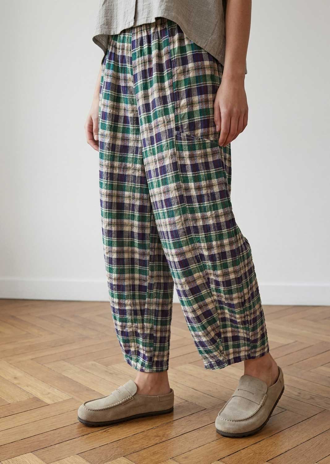 A person wearing loose, wide-leg plaid trousers in green, navy, beige, and white, paired with a light gray top and beige slip-on loafers, standing on a wooden floor against a white wall.