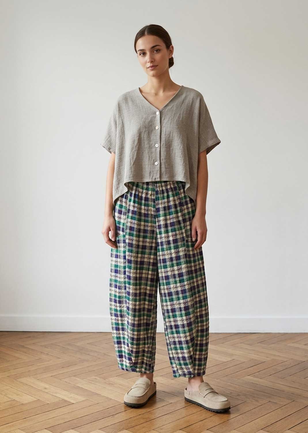 A woman stands in a minimalist room with a light gray wall and wooden parquet floor, wearing a light gray linen short-sleeve button-up shirt and green, blue, and white plaid wide-leg trousers.