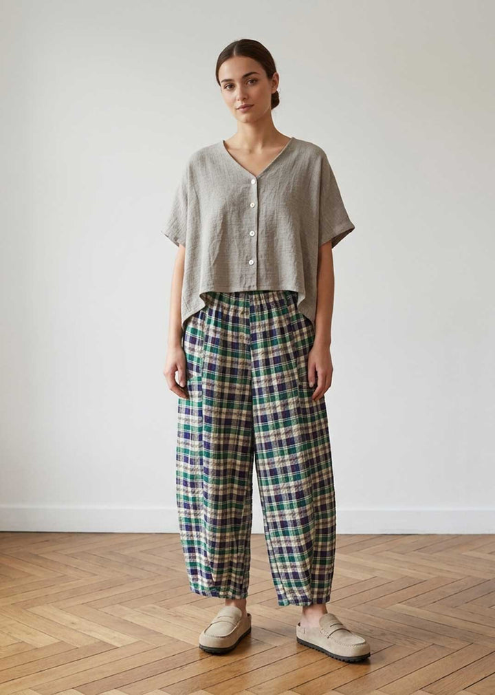 A woman stands in a minimalist room with a light gray wall and wooden parquet floor, wearing a light gray linen short-sleeve button-up shirt and green, blue, and white plaid wide-leg trousers.