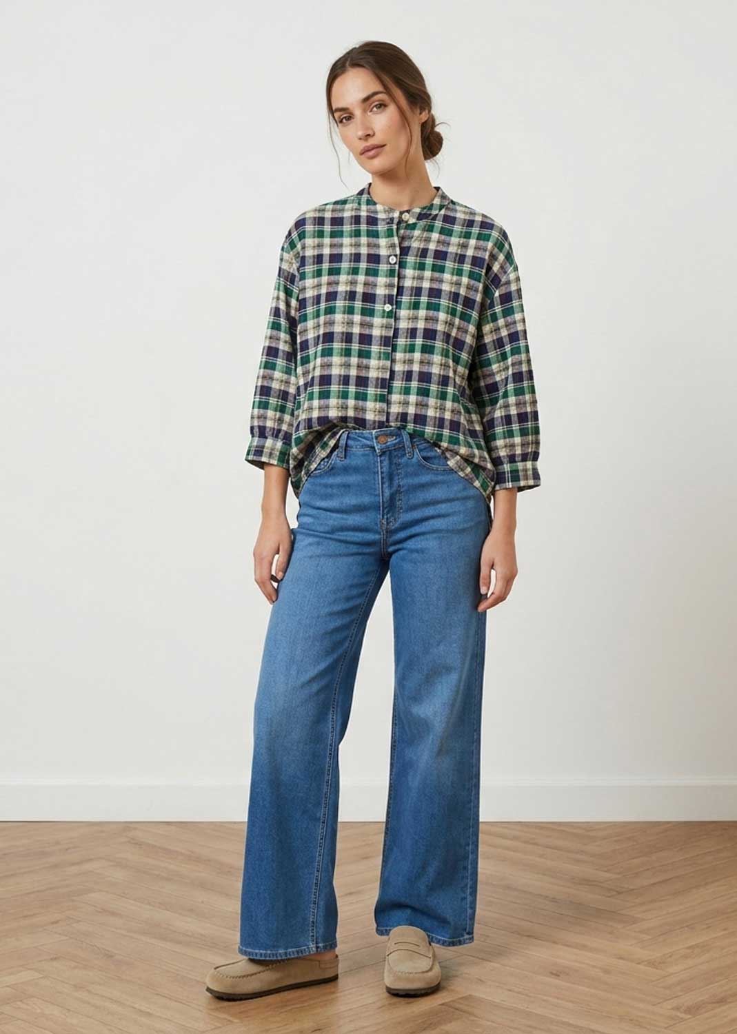 A woman modeling a green, blue, and white plaid button-up shirt with three-quarter sleeves, paired with wide-leg blue jeans and beige shoes, standing against a white wall and light wood floor.