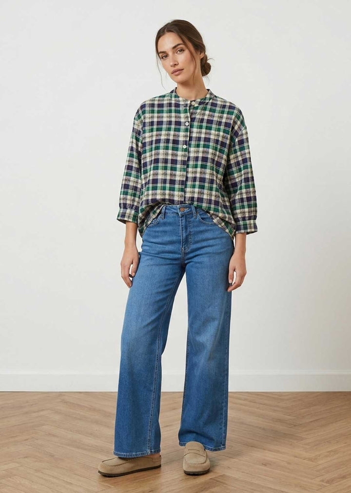 A woman modeling a green, blue, and white plaid button-up shirt with three-quarter sleeves, paired with wide-leg blue jeans and beige shoes, standing against a white wall and light wood floor.