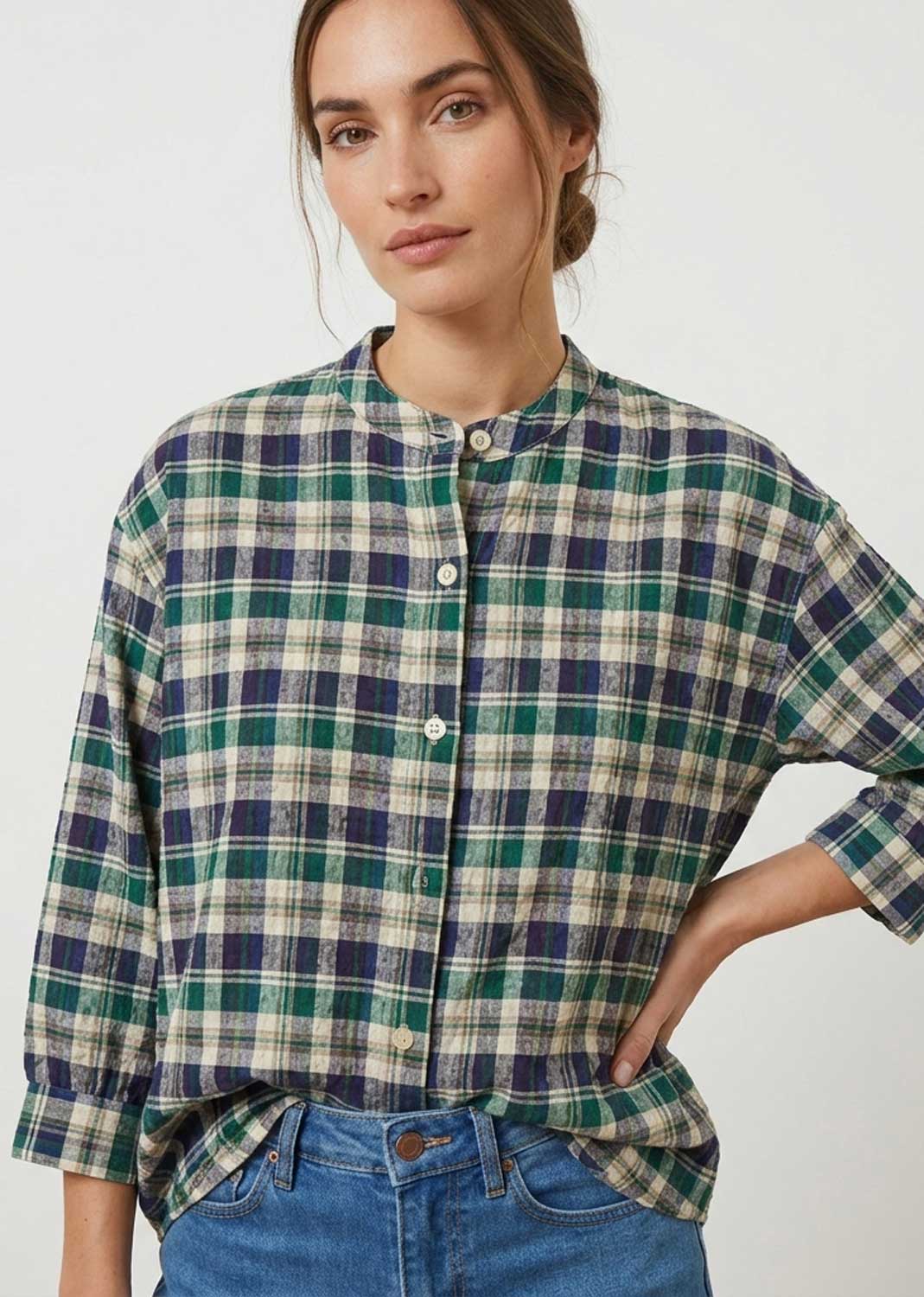 A woman wearing a green, navy, and beige plaid button-up shirt with blue jeans, photographed against a plain light gray studio background.