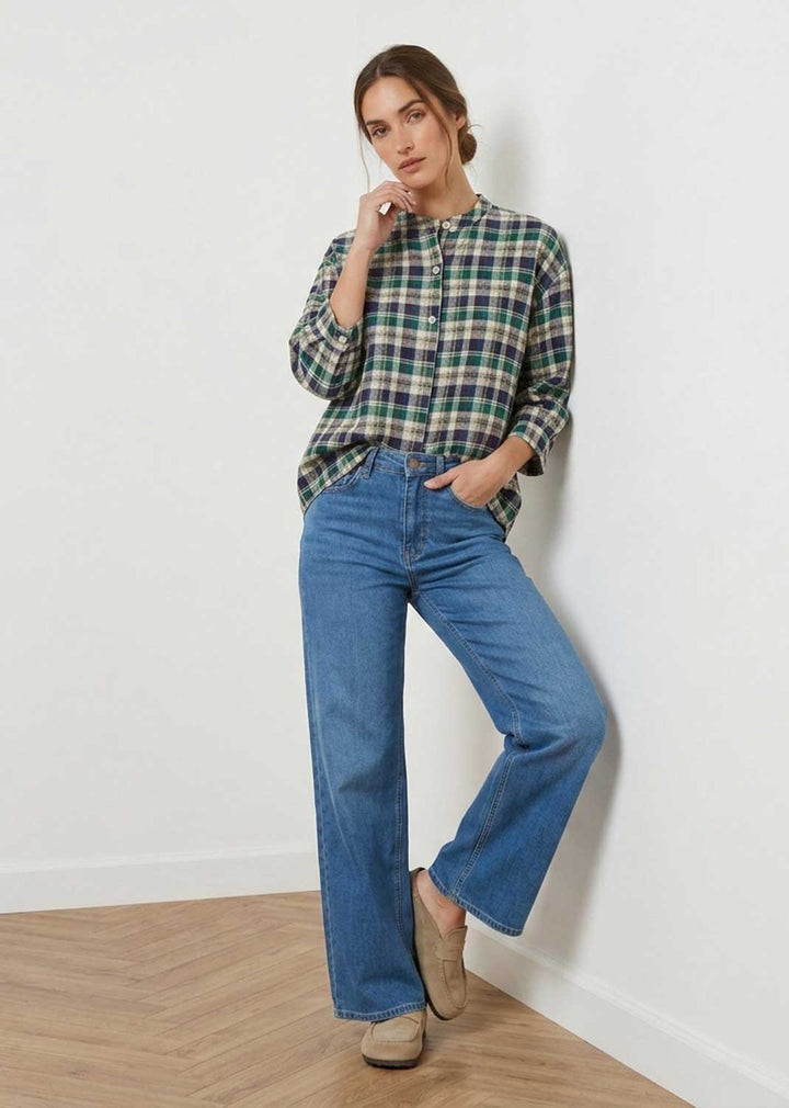A woman modeling a green and blue plaid button-down shirt with wide-leg blue jeans and beige slip-on shoes, standing against a white wall on a light wood floor.