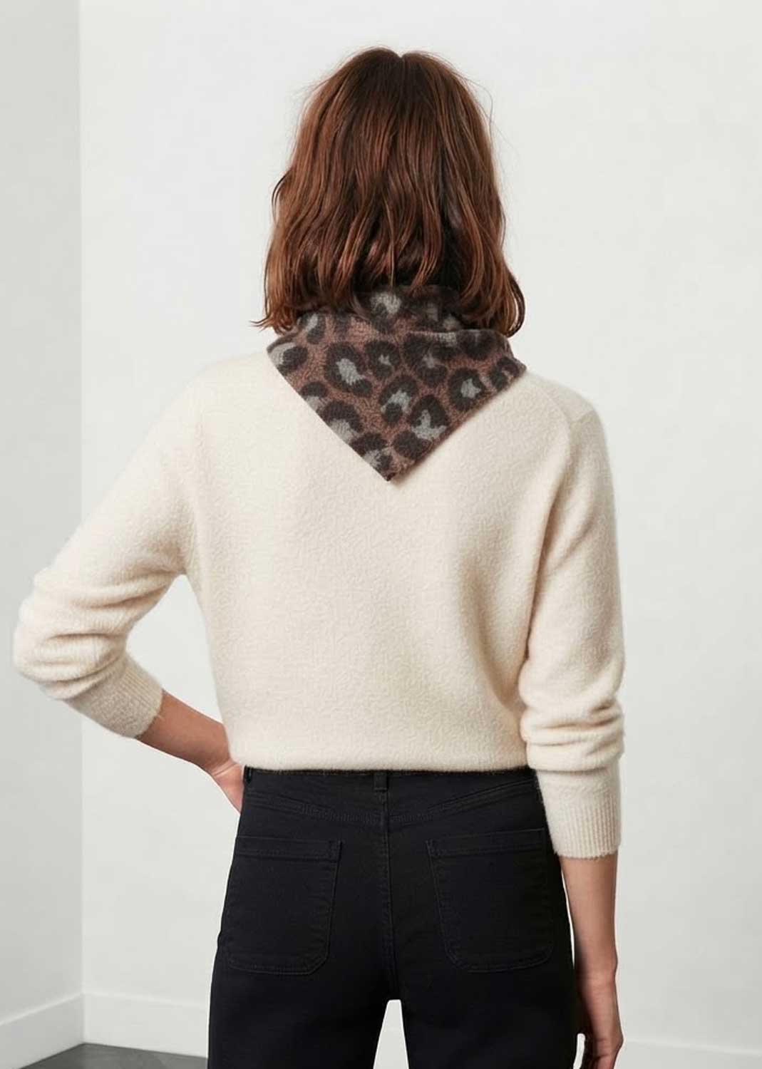 A woman viewed from the back wearing a cream-colored knit sweater with a leopard-print neck patch, paired with black pants, against a plain white wall.