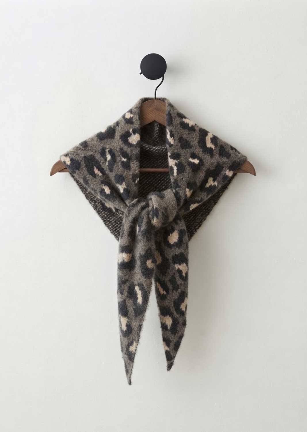 A leopard print knit scarf draped over a wooden hanger against a plain off-white wall.