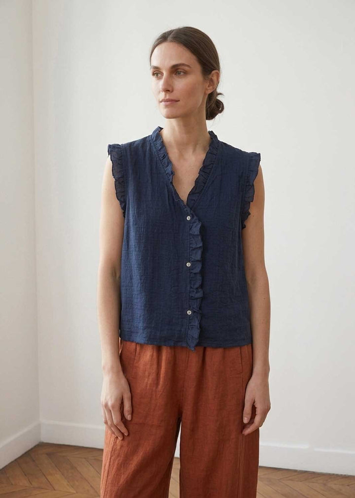 A woman wearing a sleeveless dark blue textured top with ruffle trim and rust-colored linen pants, standing in a minimalist room with white walls and a wooden floor.