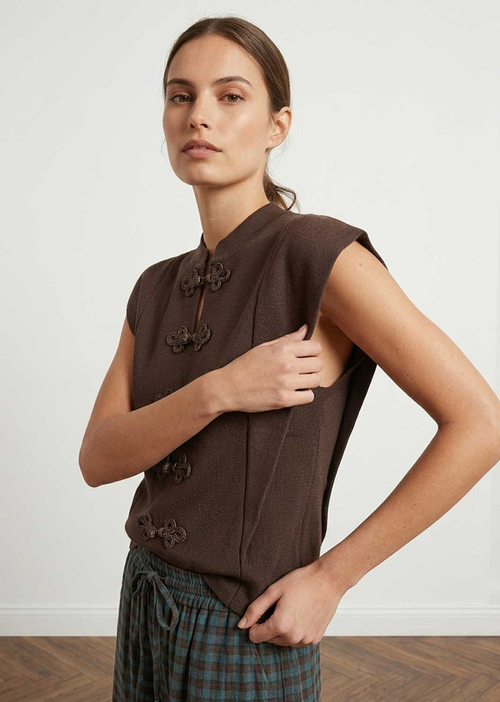 A woman modeling a sleeveless dark brown top with decorative dark brown buttons and a high collar, paired with green and blue plaid trousers, against a plain white wall.