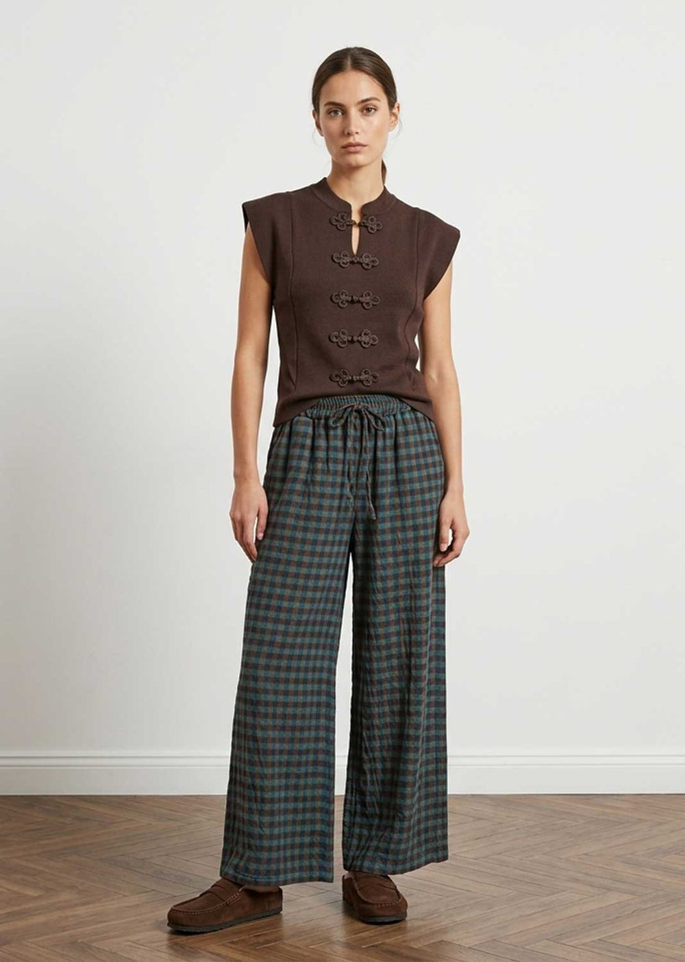 A model stands in a studio wearing a sleeveless dark brown cropped vest with front buttons and dark green and black plaid wide-leg trousers, paired with brown shoes.