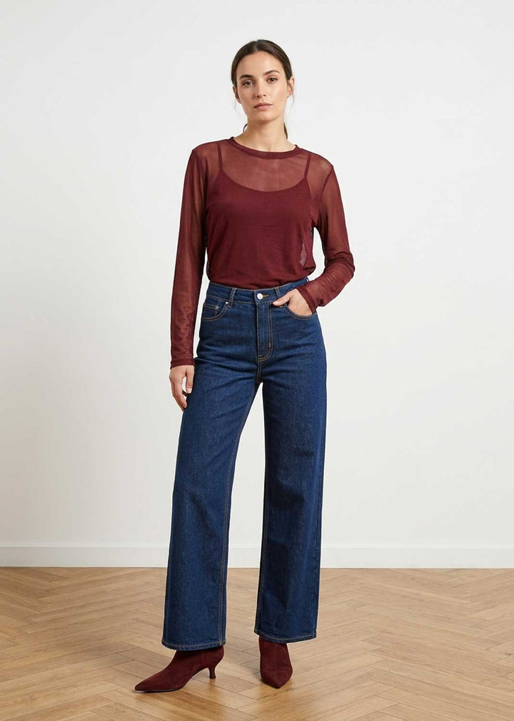A woman modeling a long-sleeve maroon sheer mesh top paired with high-waisted dark blue wide-leg jeans and burgundy heeled ankle boots, standing against a white wall on a light wood herringbone floor.