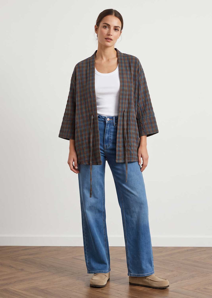 A woman stands in a studio wearing a brown and black plaid open-front cardigan over a white tank top, blue wide-leg jeans, and tan slip-on shoes against a plain light wall and wood floor.