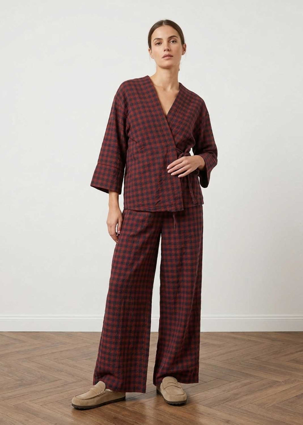 A woman modeling a matching two-piece plaid set with a wrap-style top and wide-leg pants in dark red and black, paired with beige slip-on shoes, set against a plain white wall and wood floor.