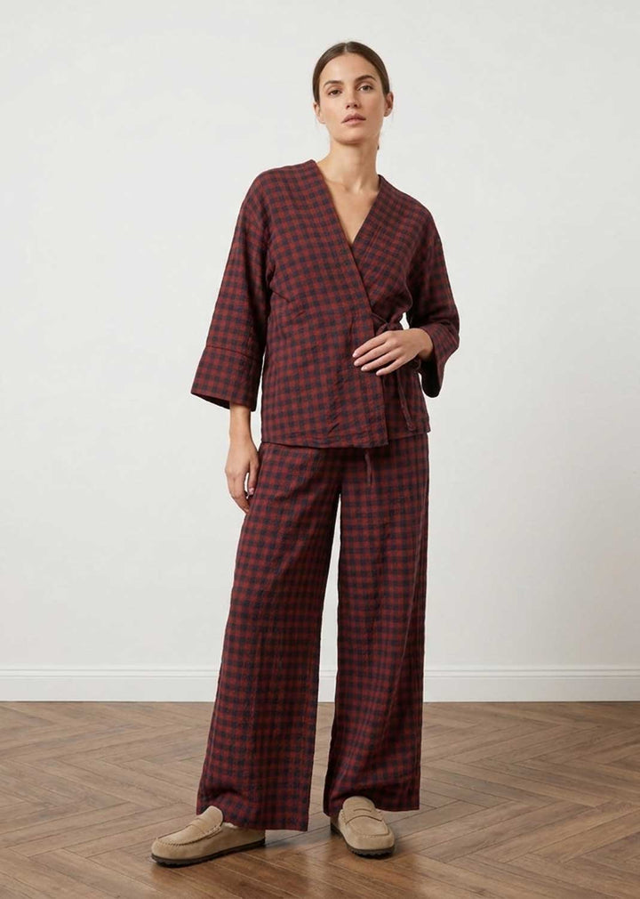 A woman modeling a matching two-piece plaid set with a wrap-style top and wide-leg pants in dark red and black, paired with beige slip-on shoes, set against a plain white wall and wood floor.