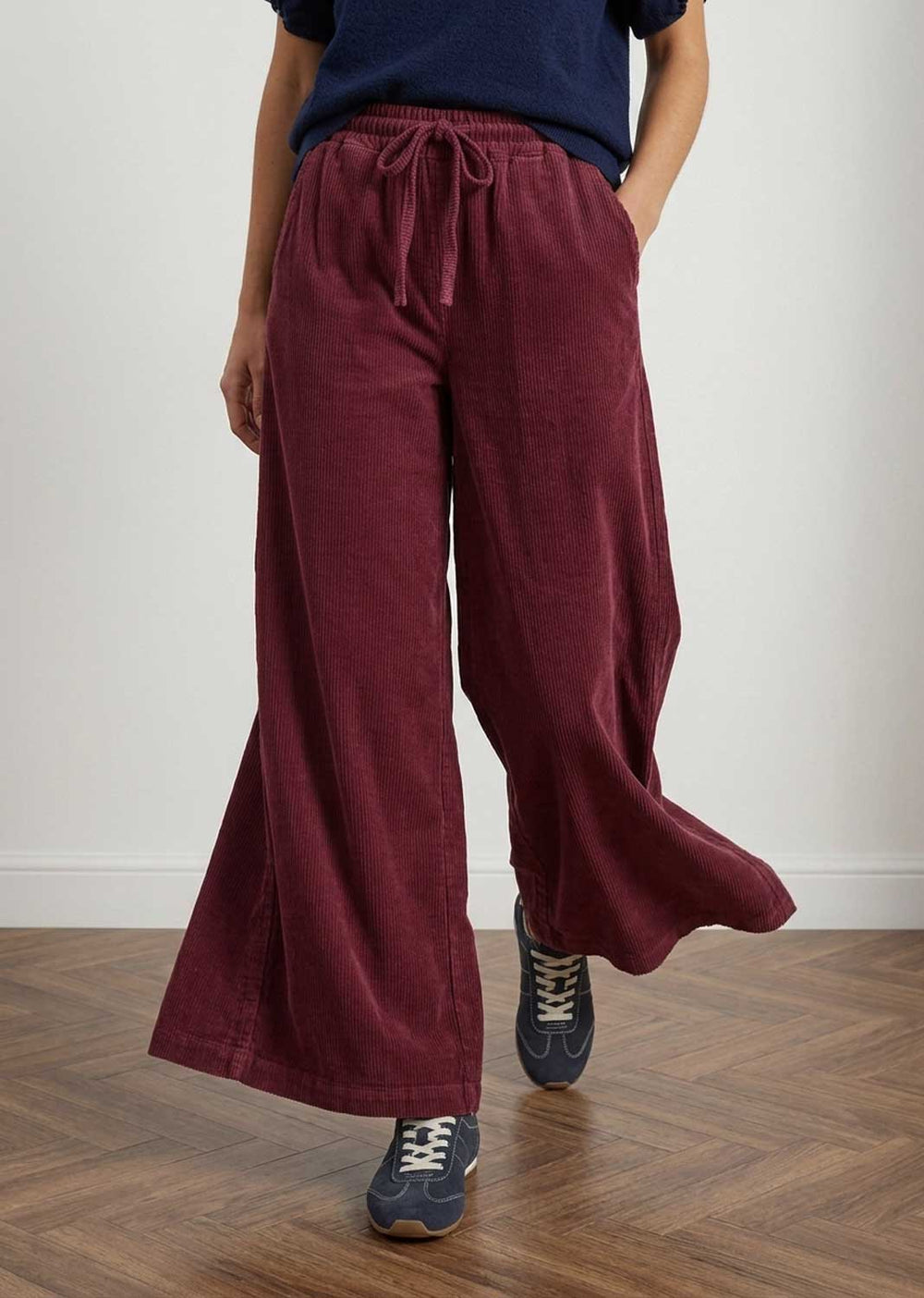 A person wearing wide-leg burgundy corduroy pants with a drawstring waist, paired with a navy top and navy sneakers, standing on a wooden floor against a plain light wall.