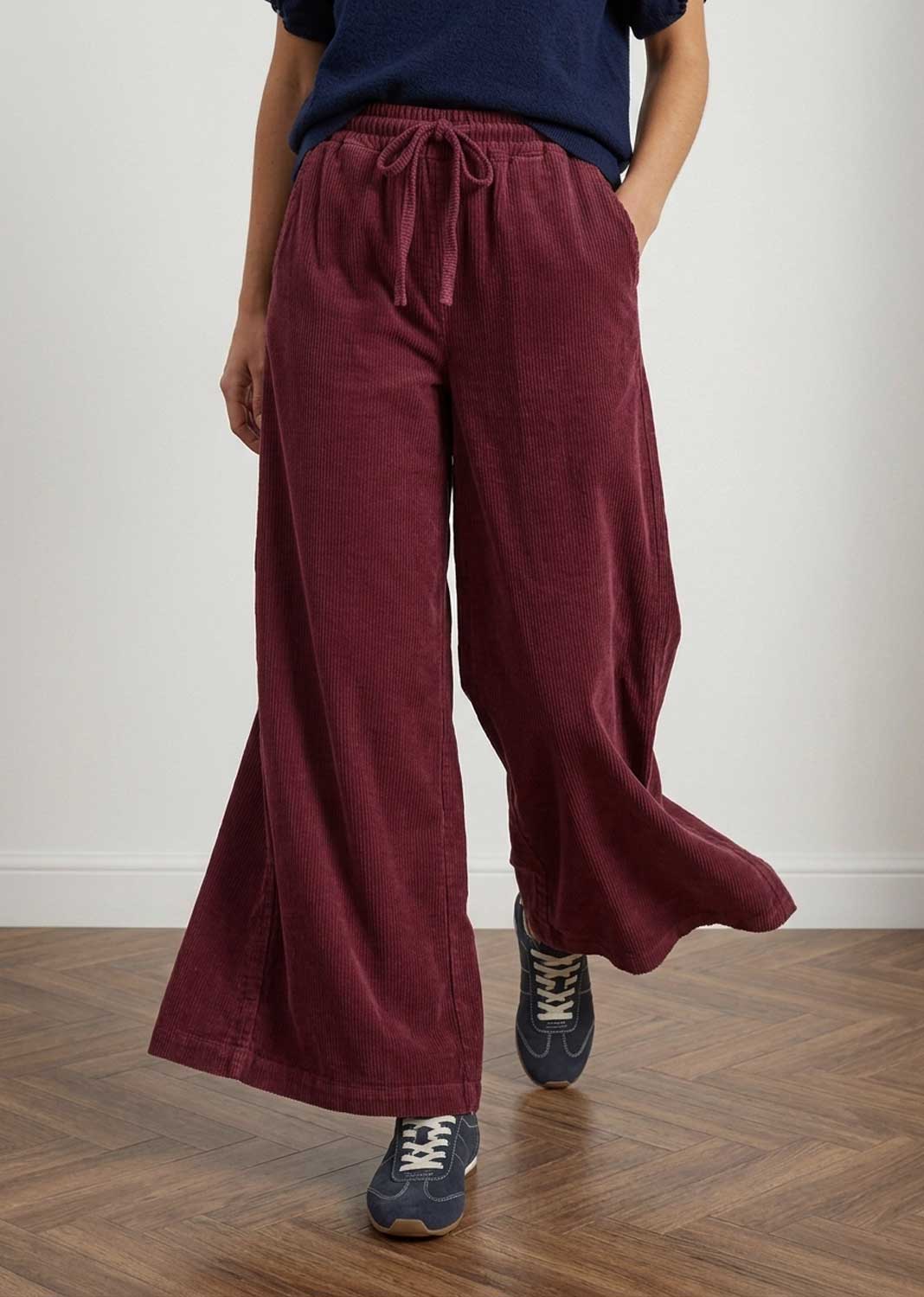 A person wearing wide-leg burgundy corduroy pants with a drawstring waist, paired with a navy top and navy sneakers, standing on a wooden floor against a plain light wall.