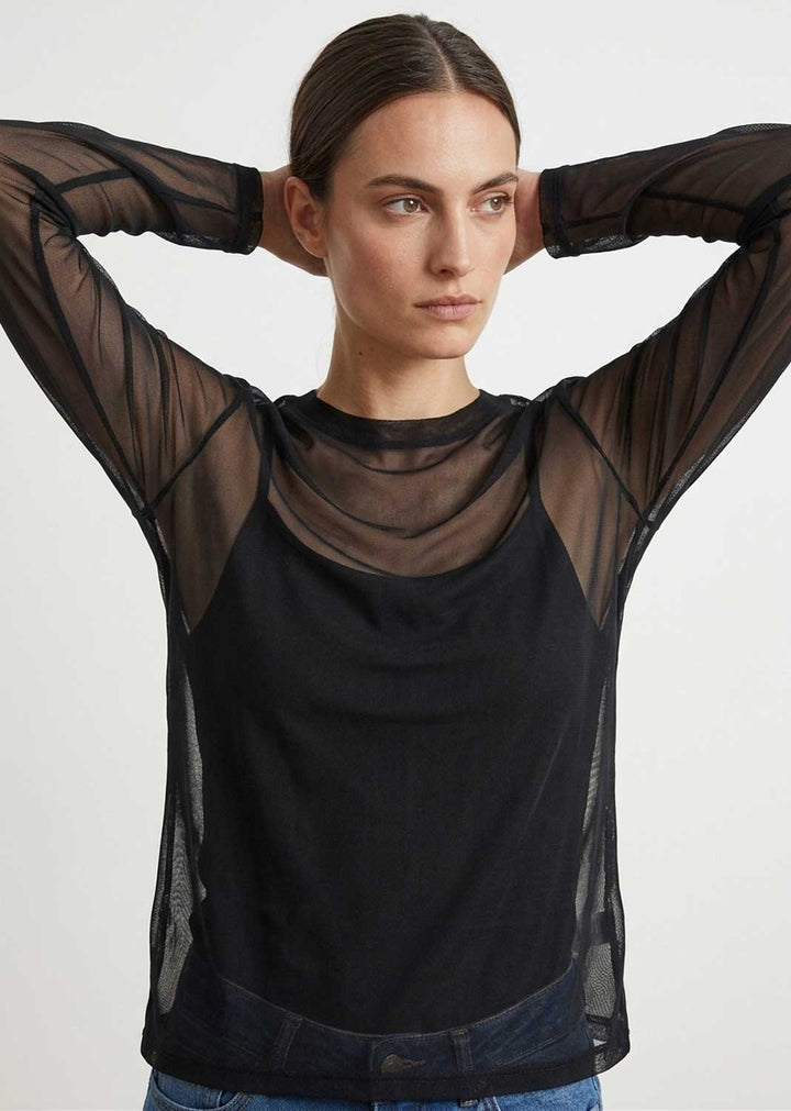 A woman wearing a black sheer mesh long-sleeve top over a black camisole, paired with blue jeans, against a plain light gray studio background.