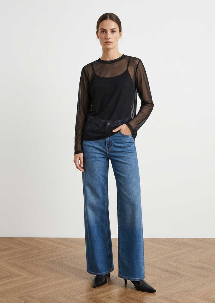 A model stands in a studio wearing a sheer black long-sleeve top over a black camisole, paired with wide-leg blue jeans and black pointed shoes, against a light gray wall and wood floor.