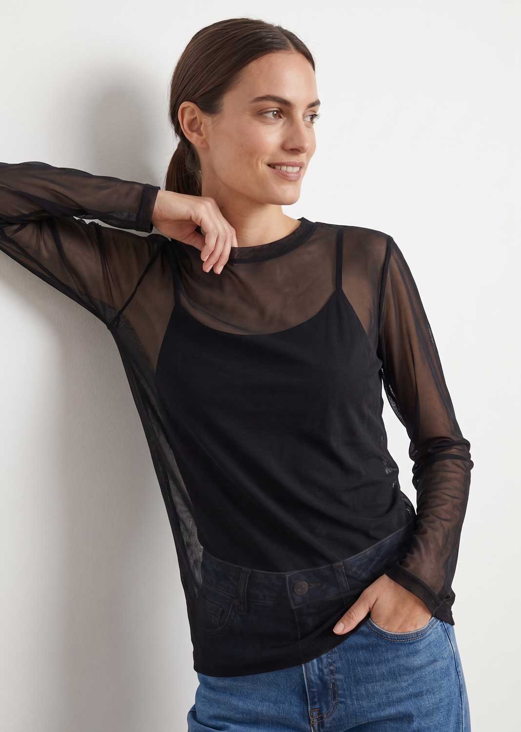 A woman wearing a black sheer long-sleeve mesh top over a black camisole, paired with blue jeans, posing against a plain white wall.
