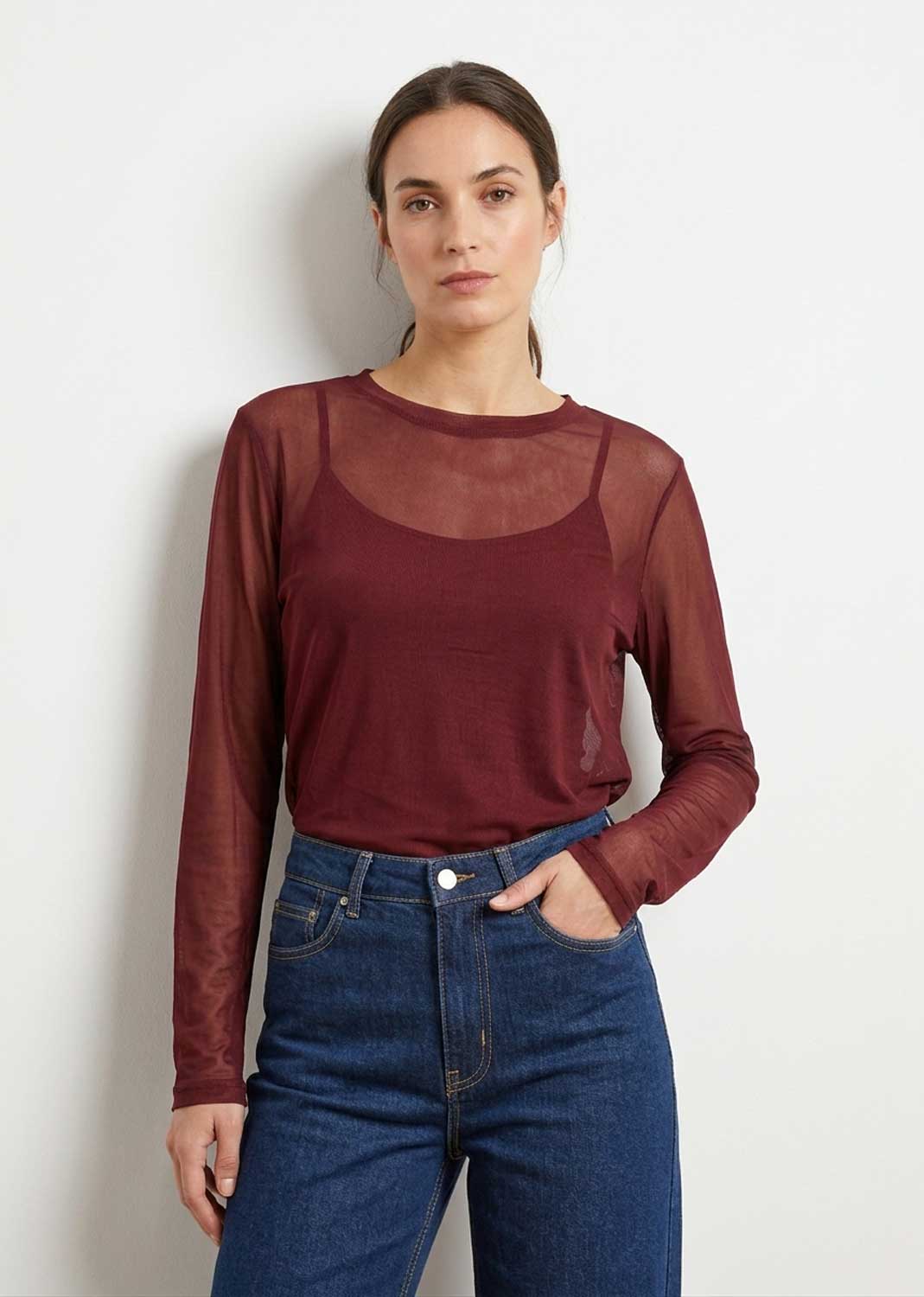A woman modeling a burgundy sheer long-sleeve top with a semi-sheer yoke over a matching solid camisole, paired with blue jeans, against a plain light gray wall.