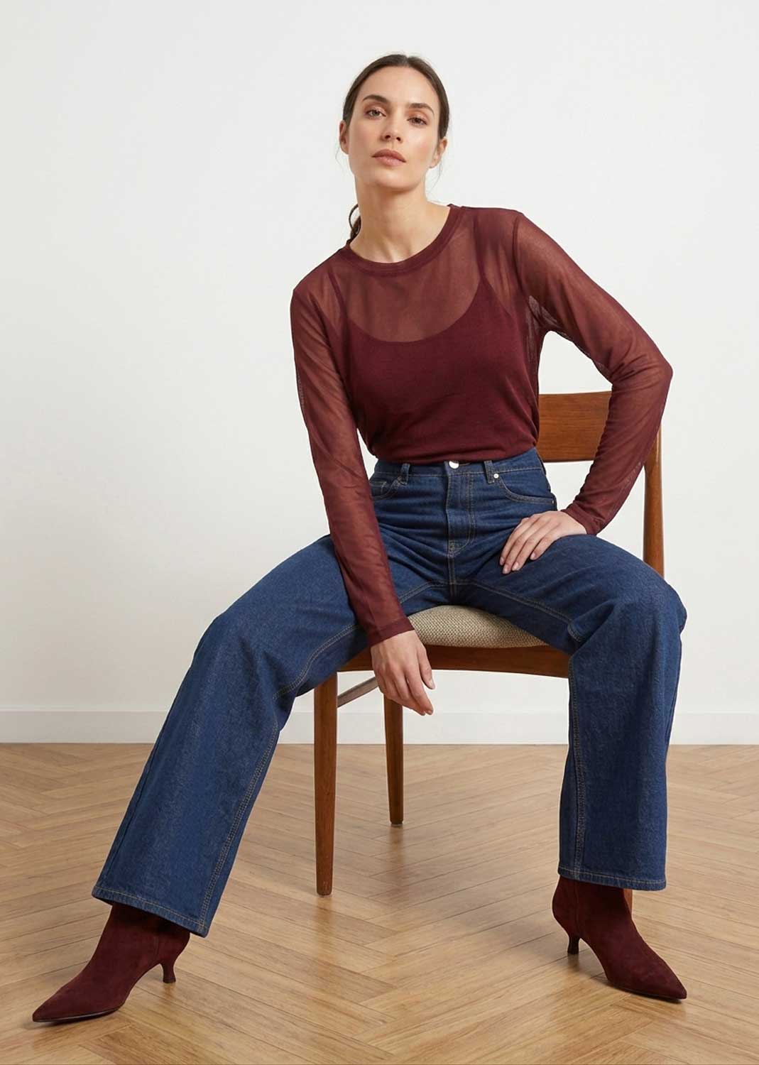 A woman sits on a wooden chair wearing a sheer burgundy long-sleeve top over a darker burgundy crewneck tee, blue jeans, and burgundy heeled ankle boots in a minimalist room with a light wall and wood floor.