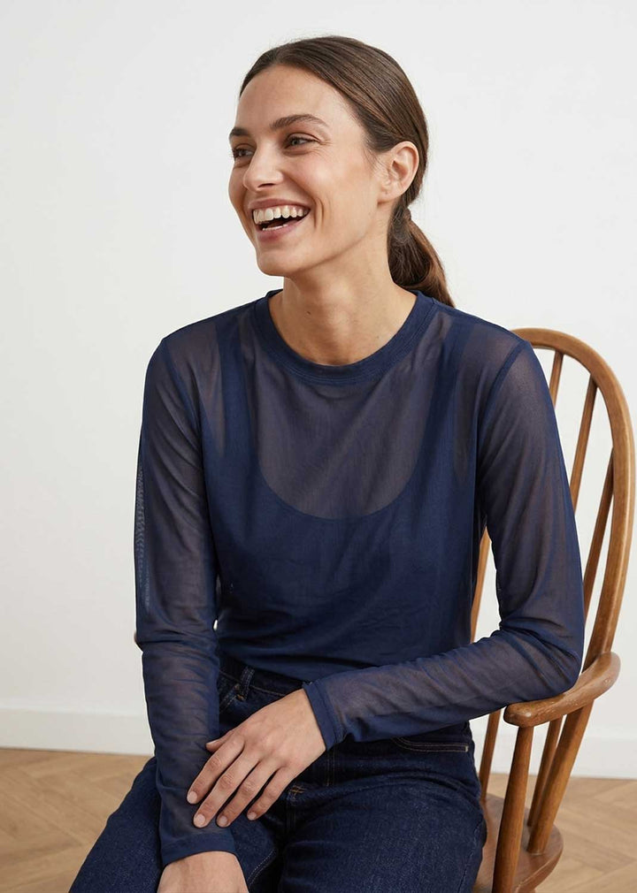 A woman smiling while wearing a navy blue long-sleeve sheer mesh top and dark jeans, seated on a wooden chair against a plain white wall.