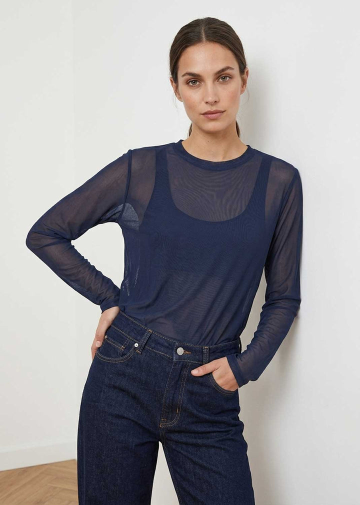 A woman modeling a navy blue sheer long-sleeve mesh top over a dark tank top, paired with dark blue jeans, against a plain white wall.