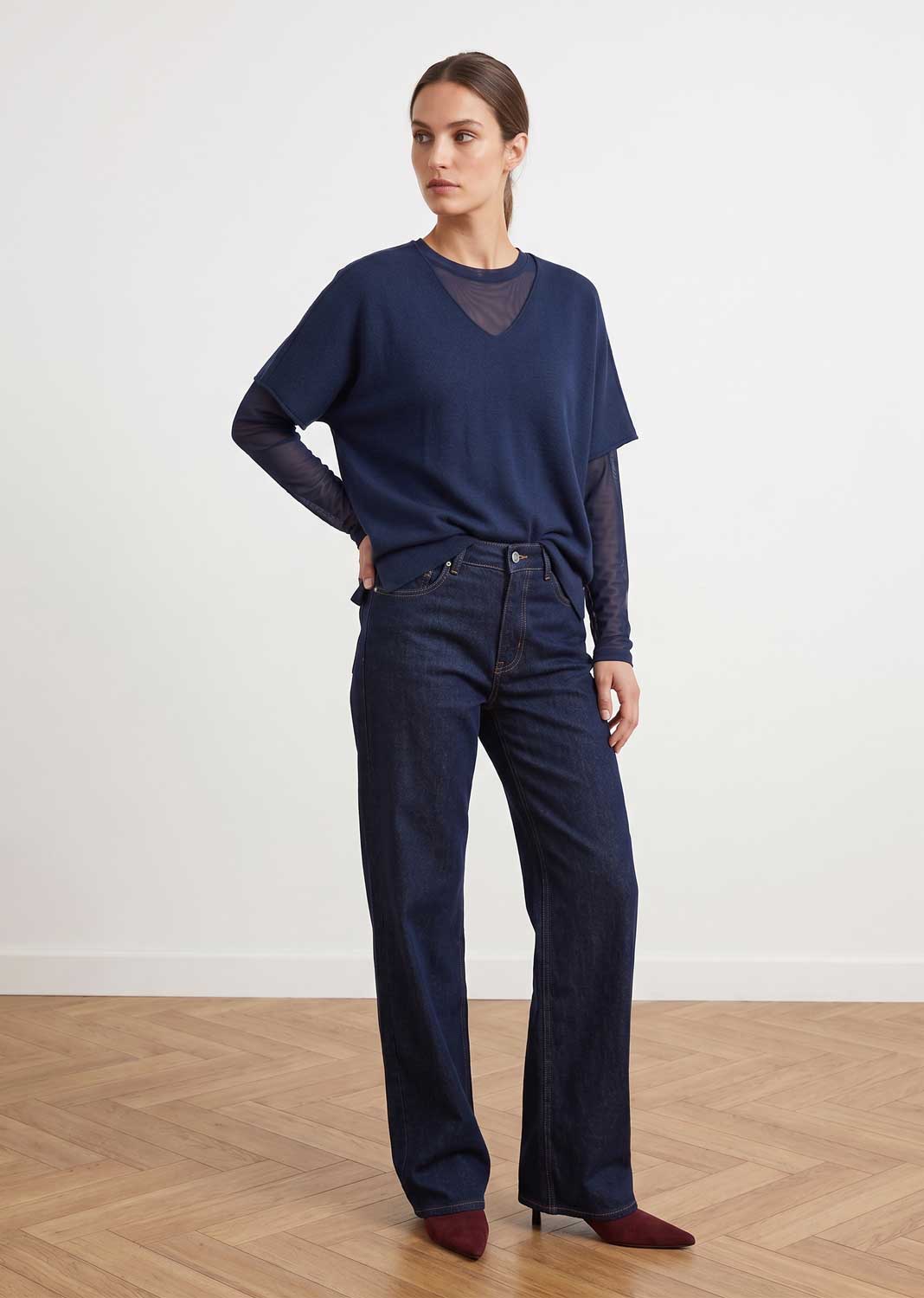 A woman modeling a navy blue knit top with sheer mesh sleeves and dark blue wide-leg jeans, standing on a light wood herringbone floor against a white wall.