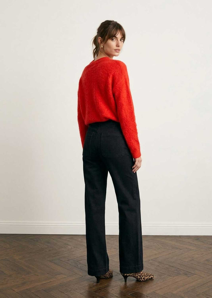 A woman stands with her back to the camera wearing a bright red fuzzy knit sweater and black wide-leg trousers, standing on a dark wood herringbone floor against a plain off-white wall.