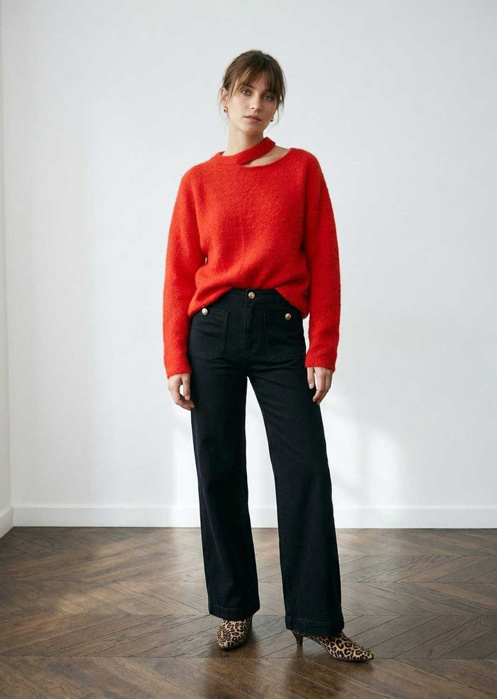 A woman stands in a bright room wearing a bright red knit sweater with a unique asymmetrical neckline, madeline black wide-leg jeans, and leopard-print pointed shoes.