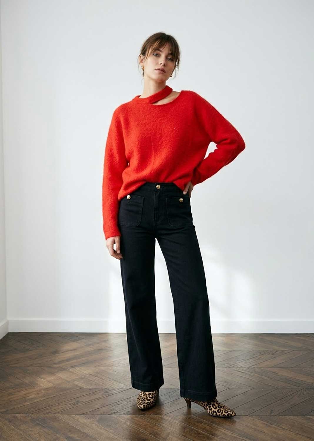 A woman modeling a bright red knit sweater with a keyhole cutout and madeline black wide-leg trousers, standing on a dark wood herringbone floor against a white wall.