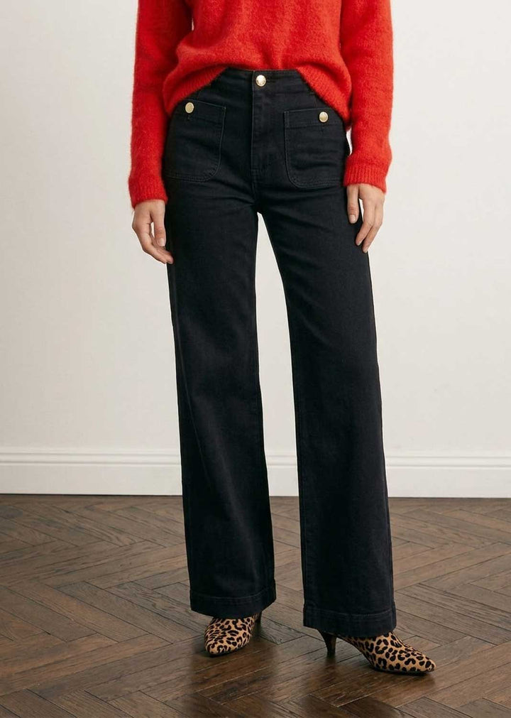 A person wearing black wide-leg jeans with gold-tone buttons, paired with a bright red sweater and leopard-print pointed heels, standing against a white wall on a wood floor.